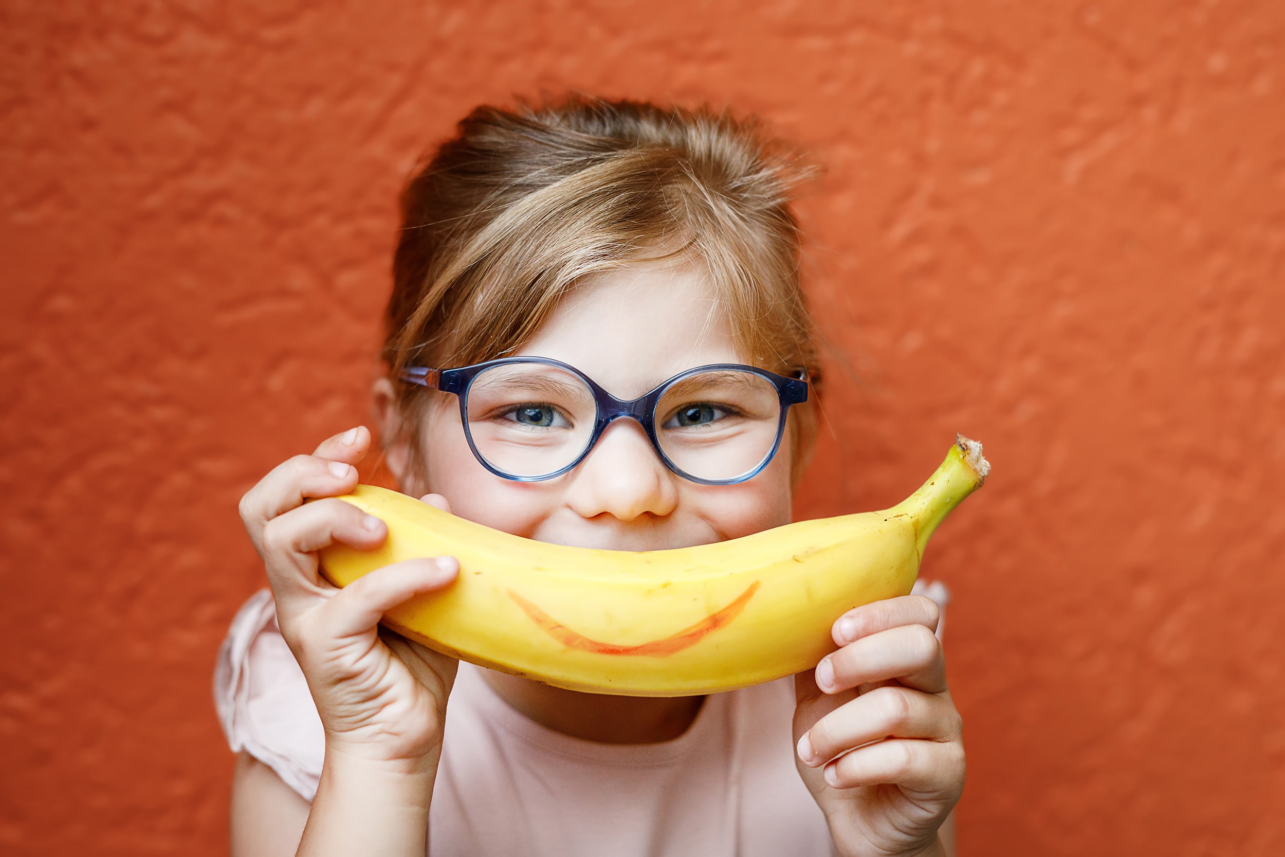 Happy little child girl with yellow banana like smile on orange background. Preschool girl with glasses smiling. Healthy fruits for children.